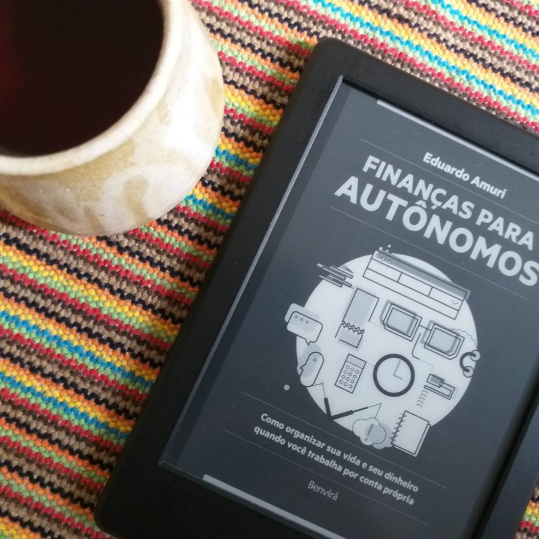 A imagem mostra um kindle com o livro 'Finanças para autônomos, de Eduardo Amuri, aberto. O kindle está sobre a mesa e há uma xícara de café ao lado.